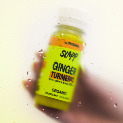 Hand holding a SLAPP Ginger Turmeric wellness shot behind foggy glass with water droplets.