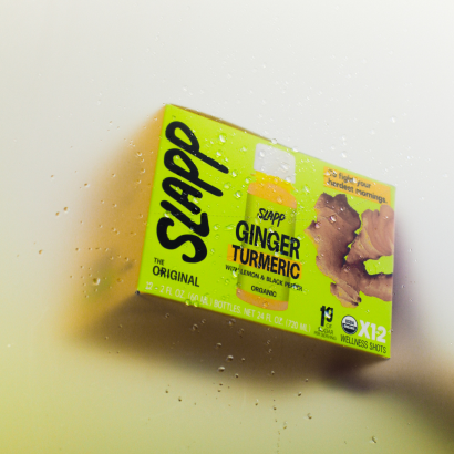 Box of SLAPP Ginger Turmeric wellness shots behind foggy glass with water droplets.